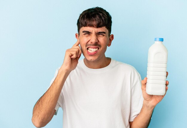 Jeune homme de race mixte tenant une bouteille de lait isolé sur fond bleu couvrant les oreilles avec les mains.