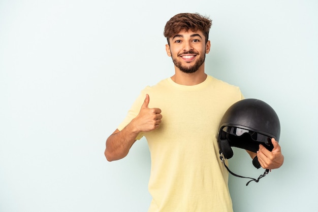 Jeune homme de race mixte tenant un casque de moto isolé sur fond bleu souriant et levant le pouce vers le haut