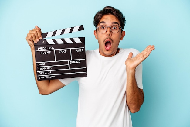 Jeune homme de race mixte tenant un clap isolé sur fond bleu surpris et choqué.