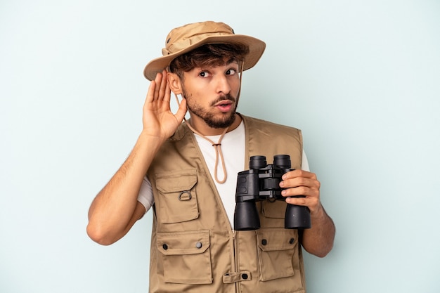 Jeune homme de race mixte tenant des jumelles isolées sur fond bleu essayant d'écouter un potin.