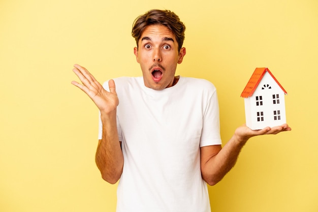 Jeune homme de race mixte tenant une maison de jouet isolée sur fond jaune surpris et choqué.