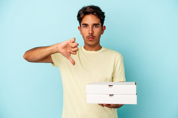 Jeune homme de race mixte tenant des pizzas isolées sur fond bleu montrant un geste d'aversion, les pouces vers le bas. Notion de désaccord.