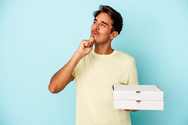 Jeune homme de race mixte tenant des pizzas isolées sur fond bleu regardant de côté avec une expression douteuse et sceptique.