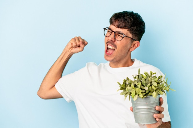 Jeune homme de race mixte tenant une plante isolée sur fond bleu levant le poing après une victoire, concept gagnant.