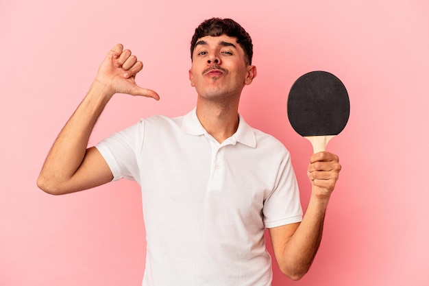 Jeune homme de race mixte tenant une raquette de ping-pong isolée sur fond rose se sent fier et confiant, exemple à suivre.