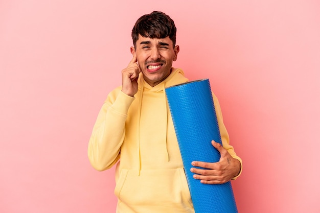 Jeune homme de race mixte tenant un tapis de sport isolé sur fond rose couvrant les oreilles avec les mains.