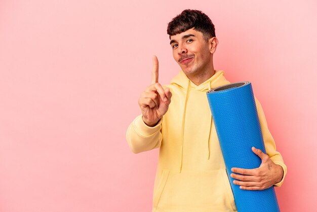 Jeune homme de race mixte tenant un tapis de sport isolé sur fond rose montrant le numéro un avec le doigt.