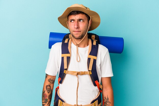 Jeune homme randonneur avec chapeau isolé sur fond bleu confus, se sent dubitatif et incertain.