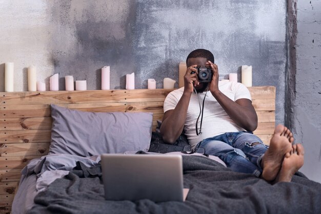 jeune homme ravi tenant un appareil photo tout en faisant des photos et couché dans la chambre.