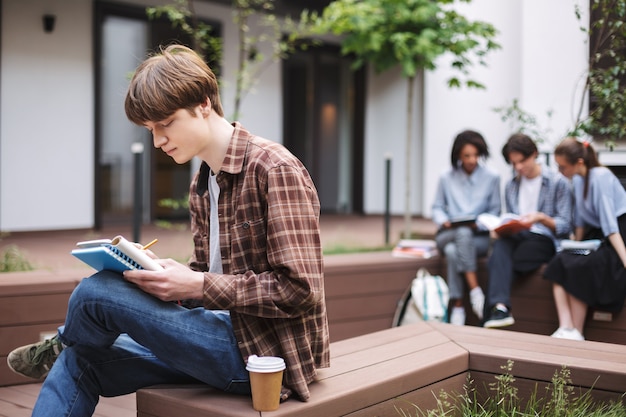 Jeune homme réfléchi assis sur un banc avec du café et de l'écriture dans un ordinateur portable tout en passant du temps dans la cour de l'université avec des étudiants en arrière-plan