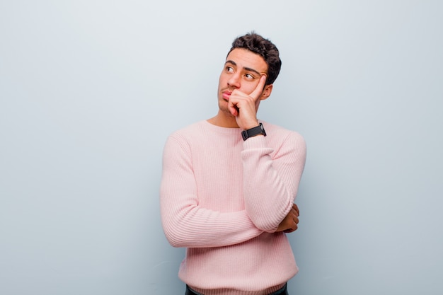 Jeune homme avec un regard concentré, se demandant avec une expression douteuse, levant les yeux et sur le côté contre le mur gris