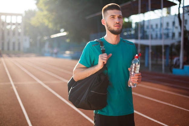 Jeune homme regardant pensivement de côté avec un sac de sport sur l'épaule et une bouteille d'eau pure à la main sur la piste de course du stade