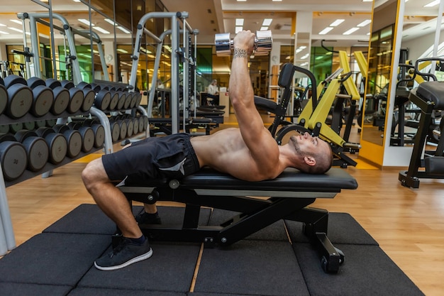 Jeune homme de remise en forme musculaire entraîne les muscles pectoraux en position couchée sur un banc avec des haltères dans les bras à la salle de sport