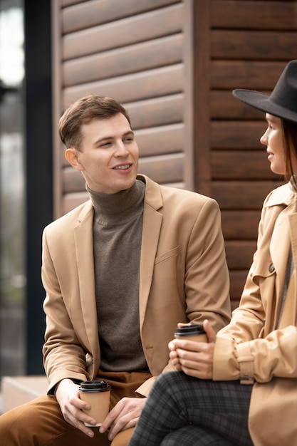 Jeune homme reposant avec un verre en regardant sa petite amie alors qu'ils étaient tous les deux assis sur un banc par l'architecture moderne et discutant pendant le froid ou le rendez-vous