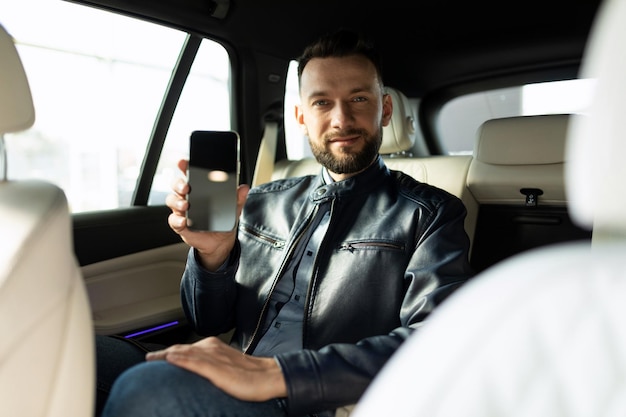 Photo le jeune homme réussi s'assied dans le siège arrière d'une voiture et démontre l'écran d'un smartphone