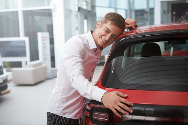 Jeune homme riant avec enthousiasme, serrant sa nouvelle automobile chez un concessionnaire automobile