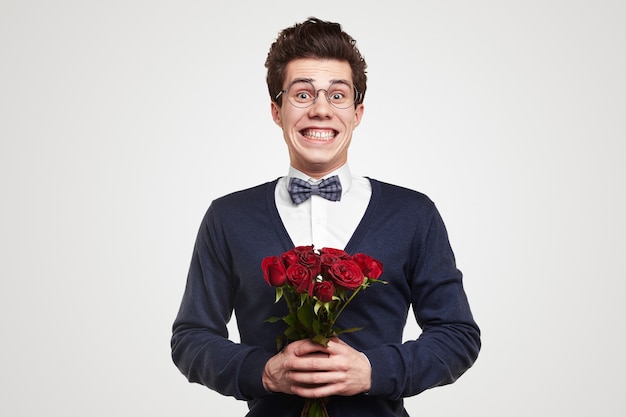 Jeune homme romantique drôle en costume élégant et lunettes ringard avec bouquet de roses rouges regardant la caméra et souriant brillamment tout en félicitant le jour de la Saint-Valentin