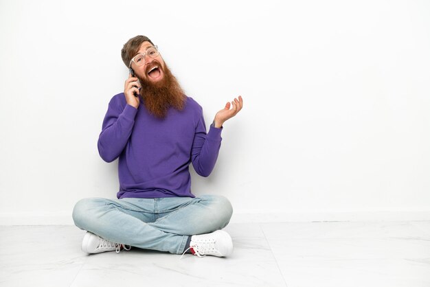 Jeune homme rougeâtre caucasien assis sur le sol isolé sur fond blanc en gardant une conversation avec le téléphone portable avec quelqu'un