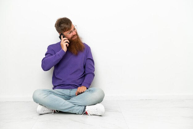 Jeune homme rougeâtre caucasien assis sur le sol isolé sur fond blanc en gardant une conversation avec le téléphone portable avec quelqu'un