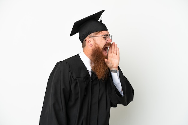 Jeune homme rougeâtre diplômé universitaire isolé sur fond blanc criant avec la bouche grande ouverte sur le côté