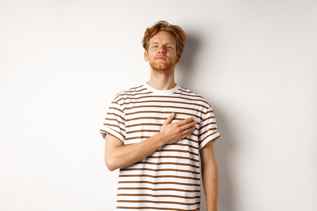 Jeune homme rousse barbu tenant la main sur le cœur et ayant l'air patriotique, debout pendant l'hymne national, debout sur fond blanc.