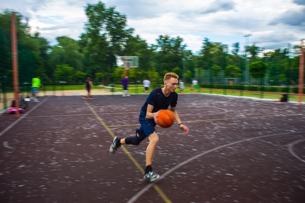 Jeune homme rousse courir et dribbler un ballon de basket à grande vitesse sur un terrain de sport dans la rue pendant la journée flou de mouvement