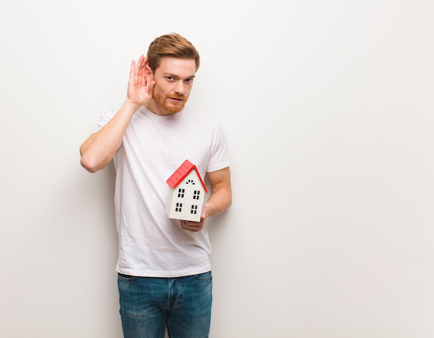 Jeune homme rousse essaie d'écouter un potin. Tenant un modèle de maison.