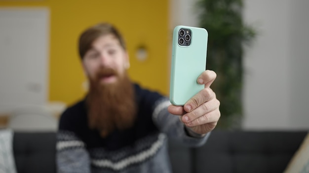 Jeune homme rousse fait selfie par smartphone assis sur un canapé à la maison