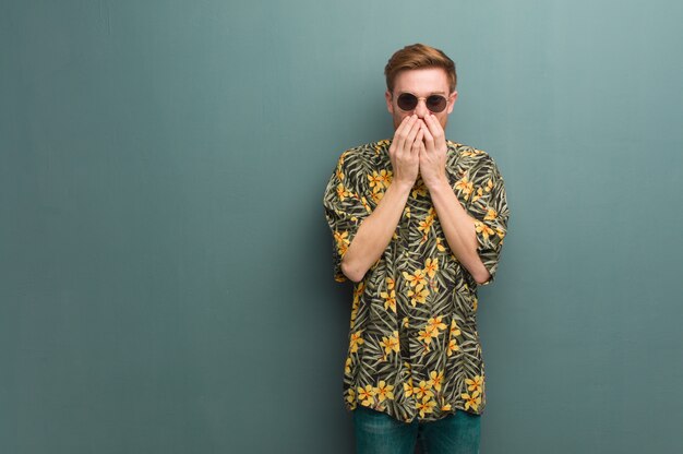 Jeune homme rousse portant des vêtements d'été exotiques rire de quelque chose, couvrant la bouche avec les mains