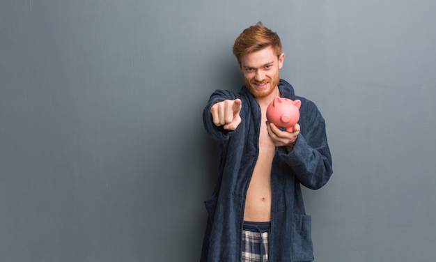 Jeune homme rousse en pyjama joyeux et pointant, tenant une tirelire.