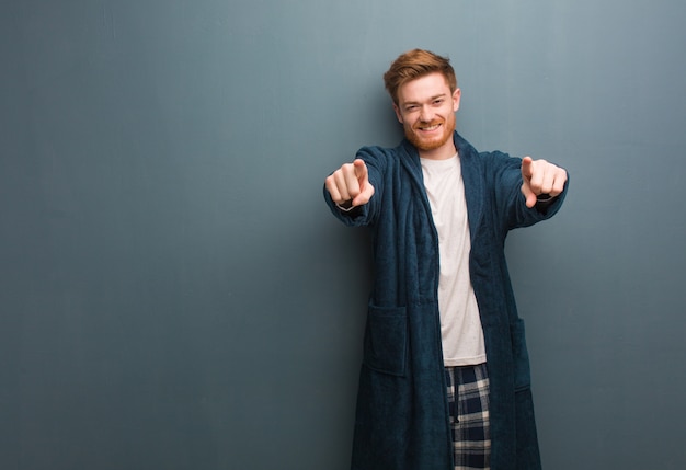 Jeune homme rousse en pyjama joyeux et souriant pointant vers l'avant