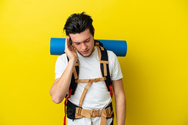 Jeune homme russe d'alpiniste avec un grand sac à dos d'isolement sur le fond jaune avec le mal de tête