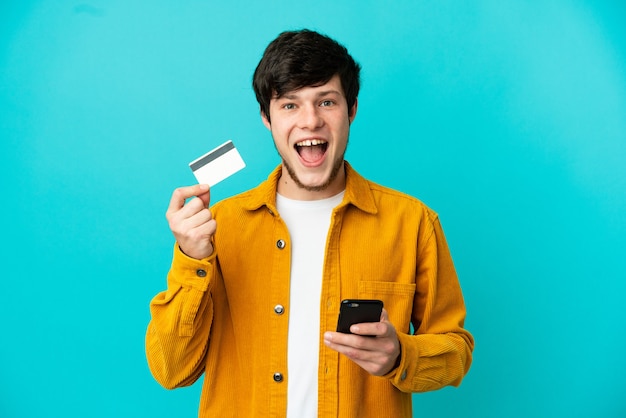 Jeune homme russe isolé sur fond bleu achetant avec le mobile et tenant une carte de crédit avec une expression surprise