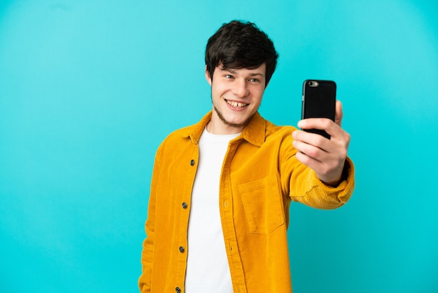 Jeune homme russe isolé sur fond bleu faisant un selfie avec téléphone portable