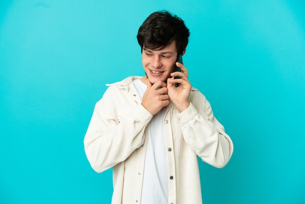 Jeune homme russe utilisant un téléphone portable isolé sur fond bleu regardant sur le côté et souriant