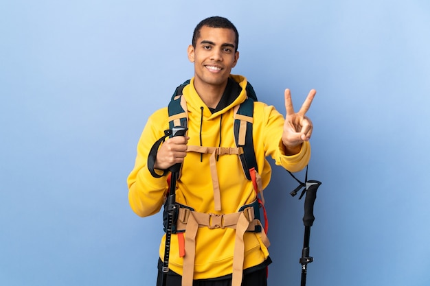 Jeune homme avec sac à dos et bâtons de randonnée sur mur isolé souriant et montrant le signe de la victoire