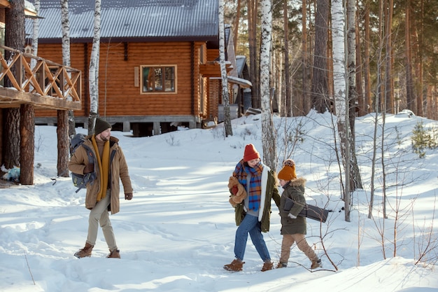 Jeune homme avec sac à dos se dirigeant vers une maison de campagne dans la forêt tandis que sa femme et leur petite fille en vêtements d'hiver chauds le suivent