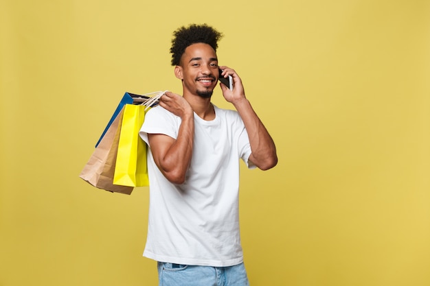 Jeune homme avec des sacs à provisions parler sur un téléphone intelligent isolé sur fond jaune