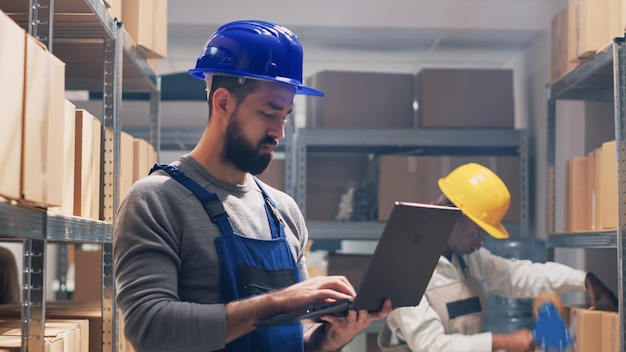 Jeune homme en salopette lisant la liste des produits dans des boîtes, planifiant la livraison pour le service industriel. Employé travaillant avec un ordinateur portable pour l'inventaire des stocks et la logistique, le développement des affaires. Prise de vue à main levée.