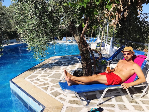 Un jeune homme sans chemise tenant un verre se détend sur une chaise de pont au bord de la piscine