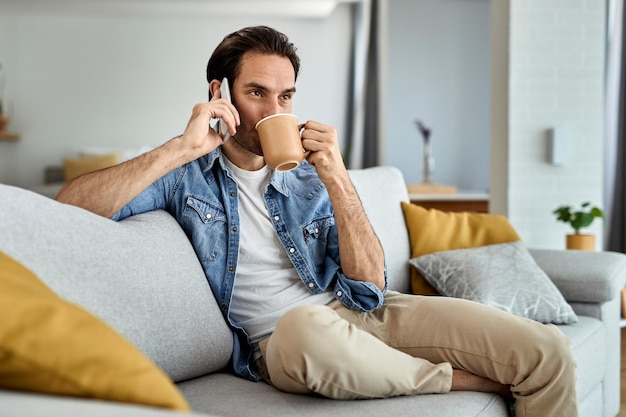 Jeune homme se détendre sur le canapé et communiquer par téléphone mobile tout en buvant du café