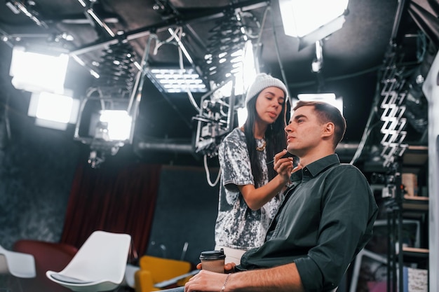Jeune homme se prépare pour la diffusion en ligne de la télévision Femme aide à se maquiller