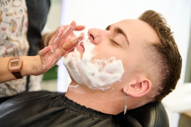 Jeune homme se rasant avec un rasoir droit par un coiffeur dans un salon de coiffure