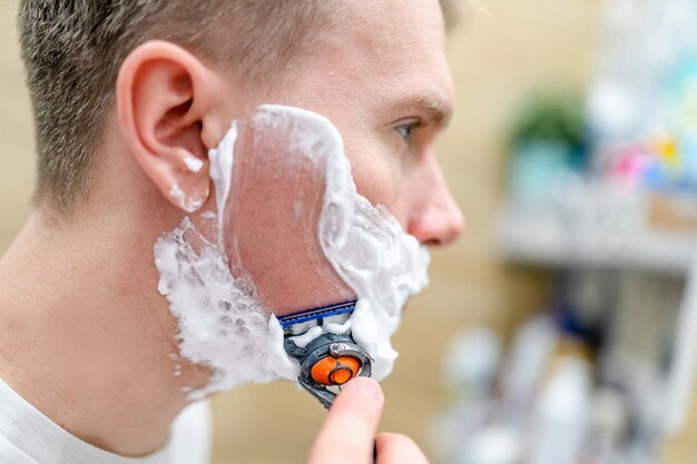 Un jeune homme se rase dans la salle de bain avec un miroir à l'aide de mousse à raser Mise au point sélective