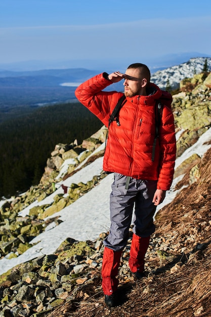 Un jeune homme se tient au sommet d'une montagne tenant une main devant ses yeux, étant protégé du soleil