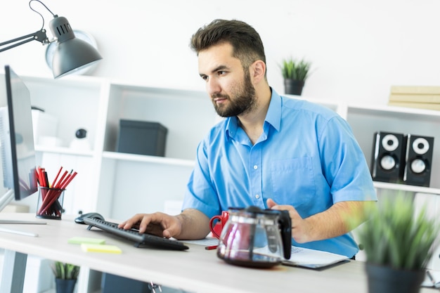 Un jeune homme se tient dans le bureau à la table d'ordinateur et prépare son café.