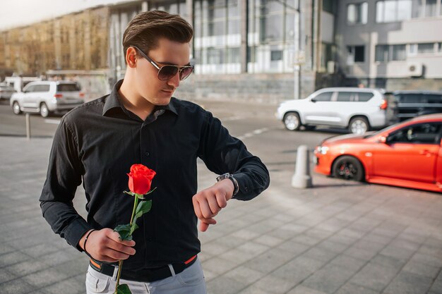 Photo jeune homme se tient à l'extérieur sur la rue et maintenez la rose rouge. il regarde les montres. guy attend. il y a une route avec des voitures derrière lui.