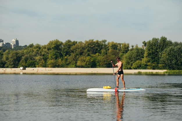 jeune homme se tient sur une planche de sup sur la rivière et pagaie sur un fond de plage et d'arbres