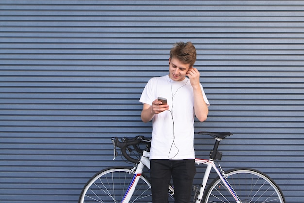 Jeune homme se tient près d'un vélo blanc écoute de la musique dans les écouteurs et regarde le smartphone