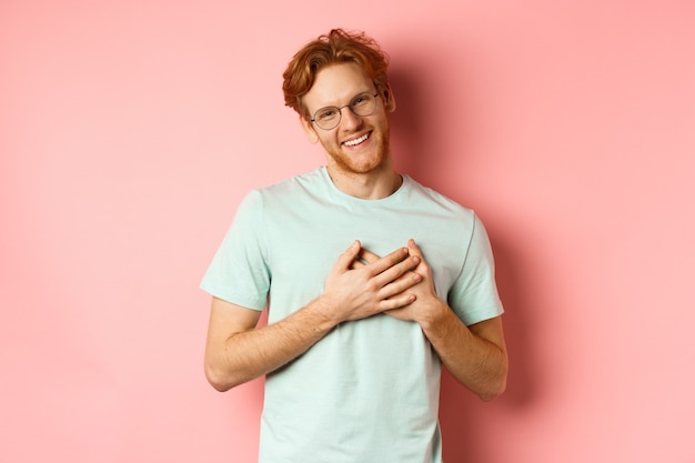 Jeune homme séduisant aux cheveux roux, tenant la main sur le cœur et souriant reconnaissant, disant merci, exprimer sa gratitude, debout sur fond rose.